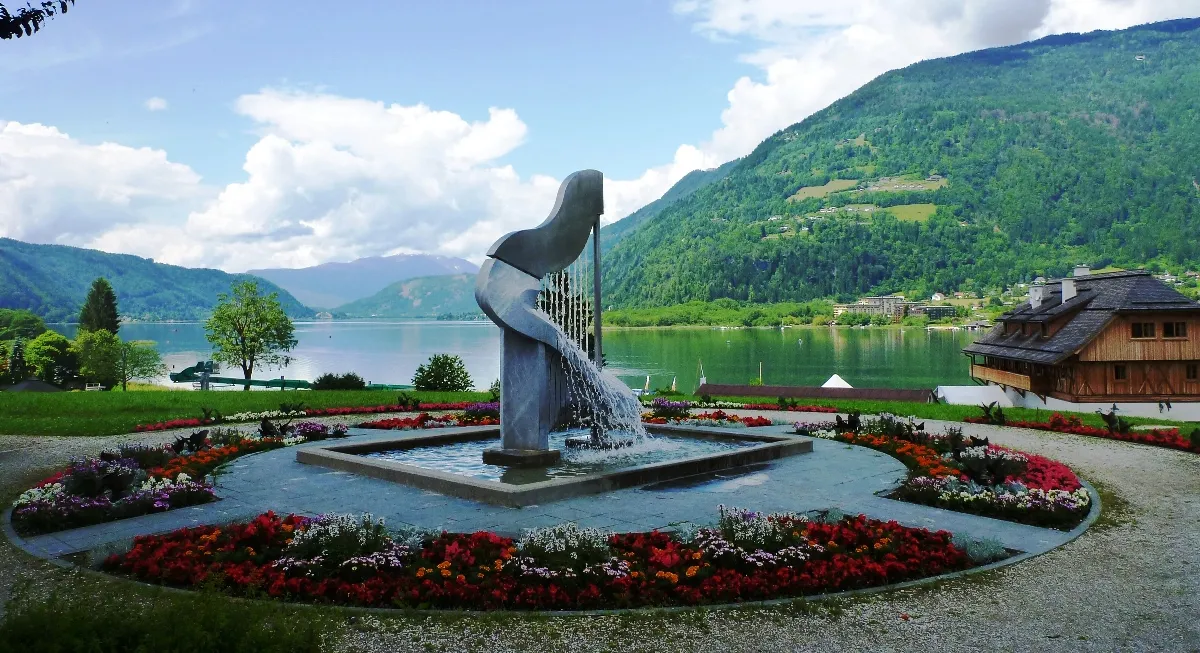 Blick über eine Parkanlage mit Blumenbeeten und einer harfenförmigen Brunnenskulptur auf den Ossiacher See und die umliegenden Berge